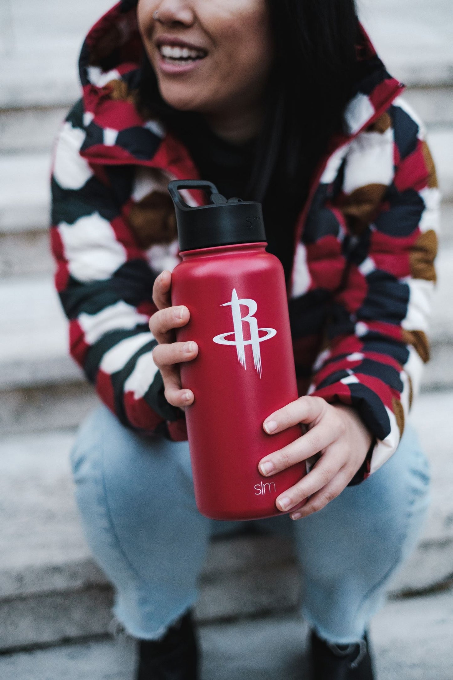 Red water bottle held in foreground with person wearing burgundy, navy blue, cream, and gold plaid jacket in background
