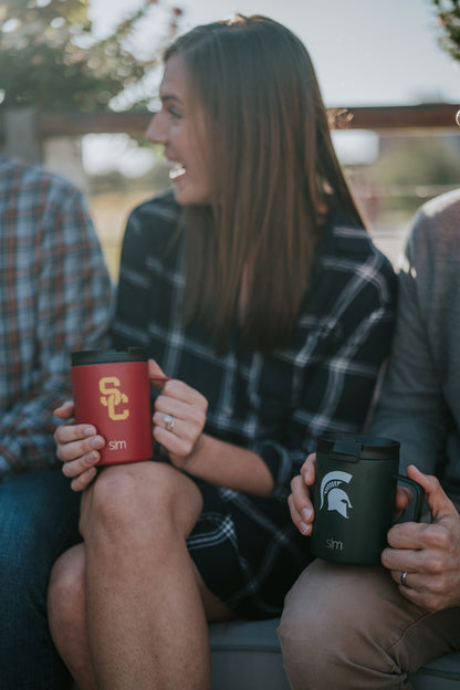 Woman in center holding bright red tumbler with gold accents, person beside her with dark forest green tumbler, warm natural tones throughout