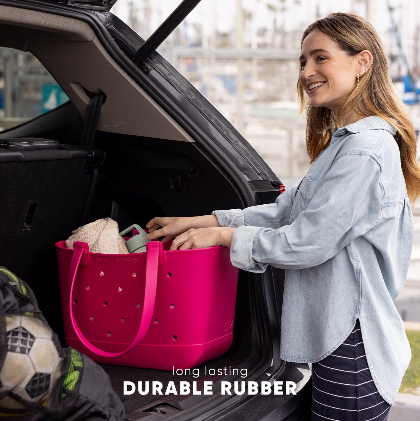 Woman placing items into a hot pink durable rubber tote bag in an open car trunk with city skyline in background. Text reads "long lasting DURABLE RUBBER."