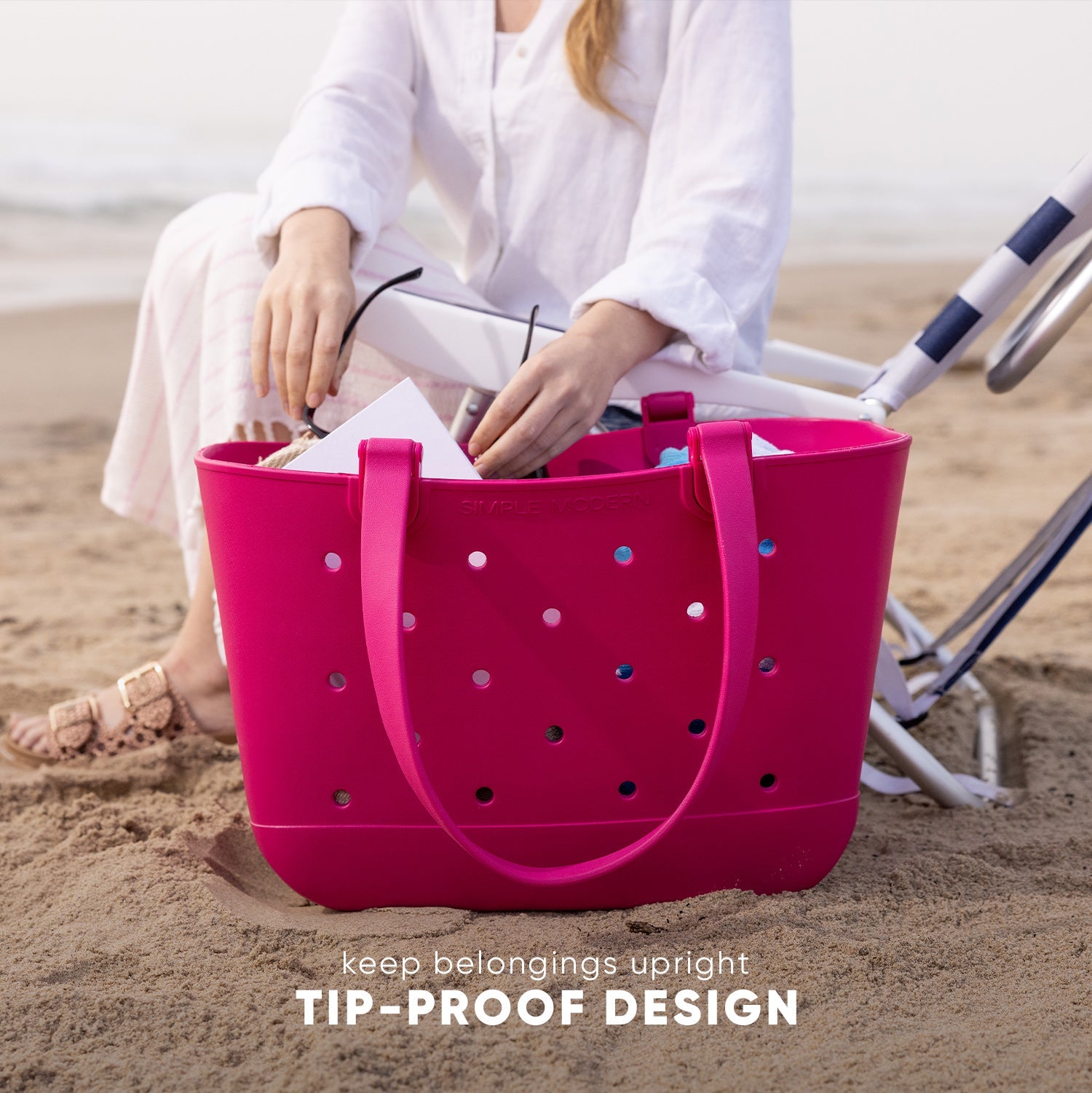 Hot pink tote bag with tip-proof perforated design held by person in white linen shirt on beach. Bag contains rolled towels and features blue, white, and black accent dots. Striped beach chair visible in background.