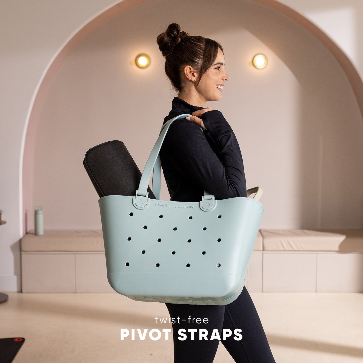 Woman in black athletic wear walking while carrying a pale blue-gray perforated tote bag with twist-free pivot straps, in a minimalist studio setting with warm lighting.