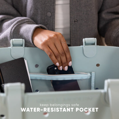 Woman placing black smartphone into water-resistant zippered pocket of light sage green Simple Modern tote bag with gray padded handles, demonstrating secure storage feature.