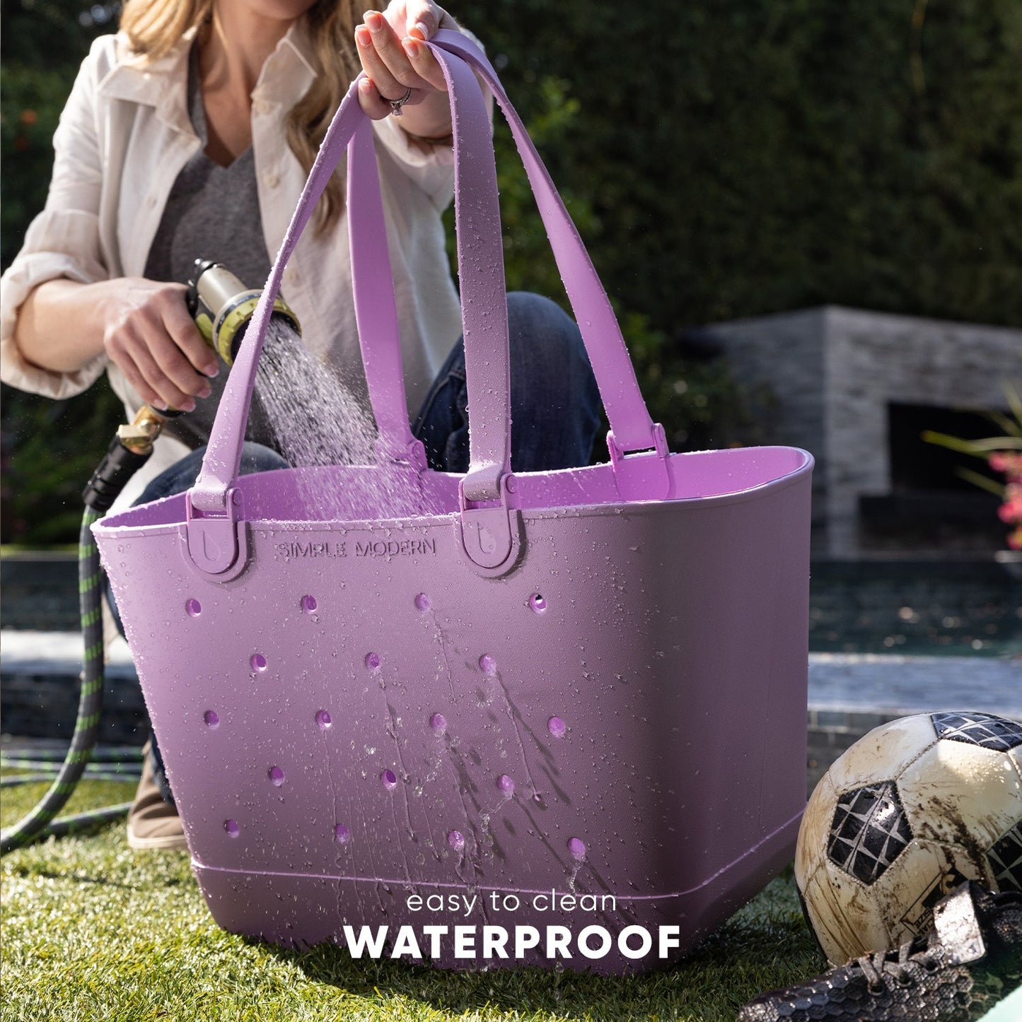 Woman holding mauve-purple Simple Modern Getaway Bag while water from garden hose sprays on it, demonstrating waterproof properties in a backyard setting with soccer ball and green lawn.